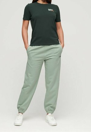Femme portant un t-shirt vert foncé à manches courtes et un pantalon de jogging vert clair avec des baskets blanches, debout les mains dans les poches.