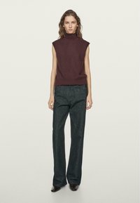 Maglione smanicato bordeaux con collo alto abbinato a pantaloni a gamba larga in denim scuro, caratterizzati da una texture liscia e un design minimale.