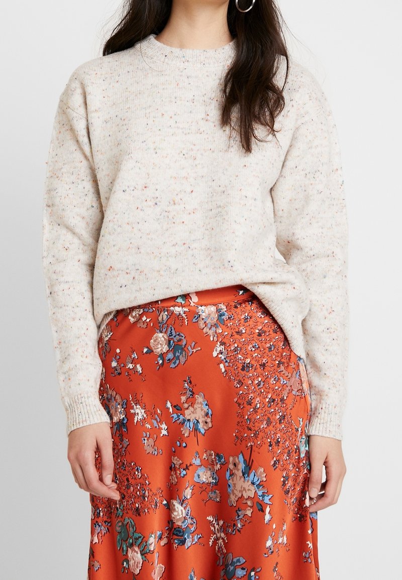 Femme portant un pull en tricot blanc moucheté associé à une jupe taille haute orange ornée de motifs floraux bleus et roses.