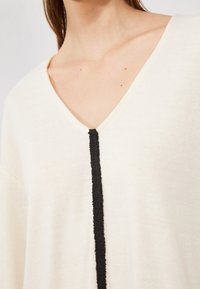 Kremfarget topp med V-hals, prydet med en vertikal svart teksturert stripe. Stoffet ser mykt og noe gjennomsiktig ut.