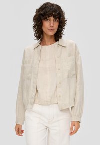 Beige linen jacket with button closure, tailored fit, two front pockets, and elasticated cuffs, paired with a matching top and light pants.