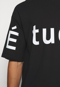 Musta lyhythihainen t-paita, jossa on suuret valkoiset tekstit. Kangas näyttää sileältä, rento istuvuus ja pyöreä pääntie.