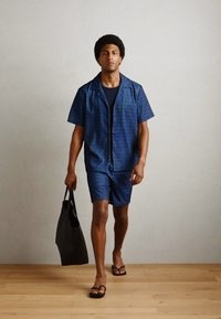 Kurzärmliges, navy-blaues Musterhemd und passende Shorts aus leichtem Material. Schwarze Tragetasche, Flip-Flops an den Füßen, neutraler Hintergrund.