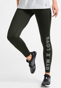 Leggings verde oliva con una texture liscia, caratterizzate dalla scritta "GYM LOVE" in bianco verticalmente lungo la gamba sinistra. Indossati con scarpe da ginnastica nere.