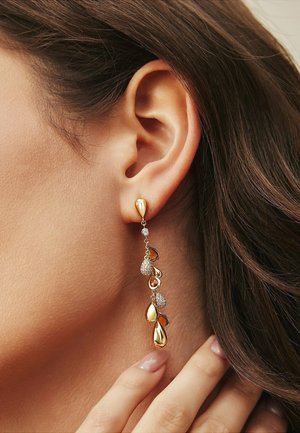 Boucles d'oreilles pendantes en ton or, avec des formes de gouttes scintillantes, reliées par une délicate chaîne en argent ornée de petits accents ronds.