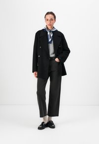 Zwarte dubbelgebreide jas met grote knopen, grijze trui, grijze hoogwaisted broek en zwarte instappers, geaccessorieerd met een blauw-witte sjaal.