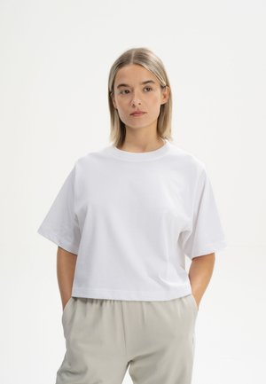 Weißes Baumwoll-Crop-Top mit kurzen Ärmeln und rundem Ausschnitt, entworfen für eine lässige Passform. Kombiniert mit hellgrauen Hosen.