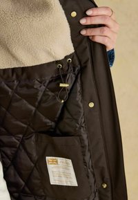 Hnedá prešívaná bunda s béžovou fleecovou podšívkou, zlatými kovovými doplnkami, detailom so šnúrkou a obdĺžnikovou tkaninovou etiketou.