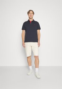 Tyrkysové polokošeľa s červenými lemami na golieri, v kombinácii s krémovými šortkami. Model nosí biele tenisky a členkové ponožky. Jednoduchý dizajn, neformálny strih.