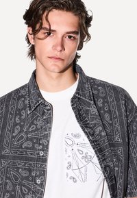 Camicia nera con motivo paisley su una t-shirt bianca che presenta un motivo simile sulla tasca. Tessuto morbido e chiusura con bottoni.