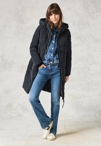 Abrigo acolchado negro con capucha, cierre de cremallera y cordones ajustables. Usado sobre un conjunto de mezclilla y zapatillas blancas, con jeans acampanados.