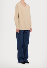 Suéter de punto acanalado beige con escote en V, combinado con jeans de mezclilla azul oscuro de corte holgado. Presenta pierna ancha y detalles de costura sutiles.