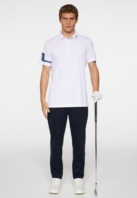 Vit skjorta med blå rand på ärmen, i kombination med marinblå byxor och vita skor. Modellen håller en golfklubba.