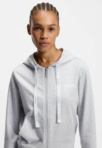 Harmaa vetoketjullinen huppari säädettävillä kiristysnauhoilla, jossa rinnan kohdalla "HUGO"-brändäys ja teksturoitu kangas, suunniteltu rentoon käyttöön.