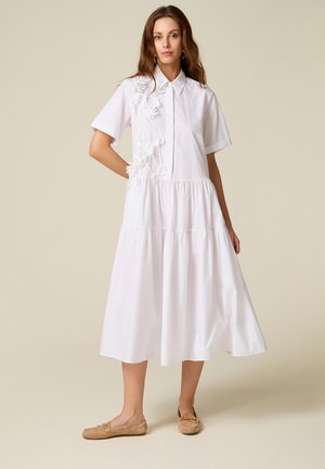 Kobieta w białej sukience midi z krótkimi rękawami, z kwiatową aplikacją na piersi oraz w beżowych mokasynach, stoi na neutralnym tle.