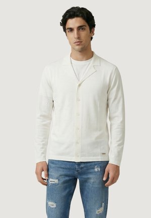 Mladý muž s tmavými vlasy, oblečený v bílém zapínaném svetru přes tričko a roztrhaných modrých džínách, stojící na světlém pozadí.