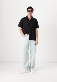 Chemise noire à manches courtes avec une texture à motif, jean large bleu clair, et baskets blanches, complétées par des lunettes de soleil noires.