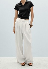 Polo noir à manches courtes, associé à un pantalon blanc ample taille haute ; des bracelets bangles marron et des sandales plates complètent le look.