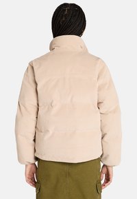 Giacca puffer in corduroy beige con colletto alto, caratterizzata da una superficie testurizzata e una silhouette leggermente imbottita. Indossata con un fondo verde oliva.