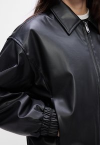 Veste noire en cuir synthétique avec fermeture éclair à l'avant, large col et poignets élastiqués, portée sur un top blanc.