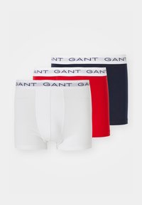 Trois paires de boxers pour hommes en blanc, rouge et marine, chacune avec une ceinture élastique blanche marquée "GANT" en bleu foncé.