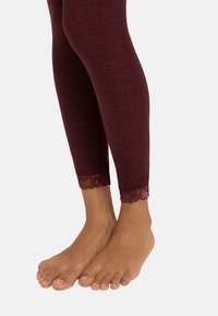 Bordeaux leggings met ribbeltextuur, afgewerkt met een kantdetails bij de enkel. Close-up van voeten die op een lichte achtergrond staan.