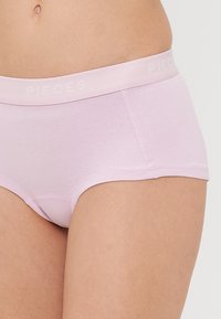 Ροζ βαμβακερό σλιπάκι με μαλακή ελαστική ζώνη που φέρει το λογότυπο "PIECES" σε λευκό χρώμα. Ασύμμετρη σχεδίαση με μέτρια κάλυψη.