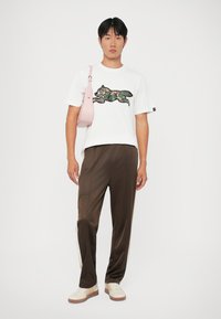 Bílý tričko s camo grafikou, hnědé tepláky s bílými bočními pruhy, světle růžová crossbody taška a béžové tenisky se světle hnědými podrážkami.