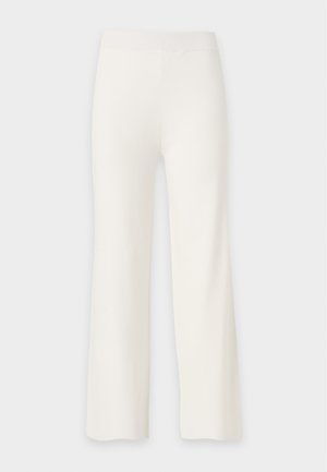 Visoka osoba tamne kože nosi bijelu top s izrezima dugih rukava, široke krem pantalone i crne šiljate cipele, stoji na jednobojnoj pozadini.