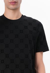 T-shirt noir avec un motif géométrique en relief, manches courtes, col rond et texture douce. Convient pour une tenue décontractée.