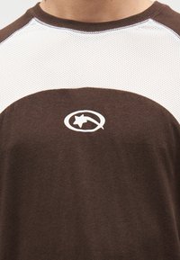 Καφέ και λευκό κοντομάνικο πουκάμισο με στρογγυλή λαιμόκοψη, ύφασμα από δίχτυ στα μανίκια και λ ογότυπο με αστέρι τυπωμένο στο μπροστινό μέρος.