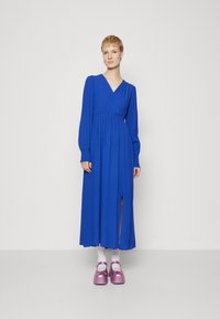 Abito blu a maniche lunghe con design avvolgente, tessuto testurizzato e uno spacco fino alla coscia. Indossato con calzini bianchi e scarpe con piattaforma rosa.