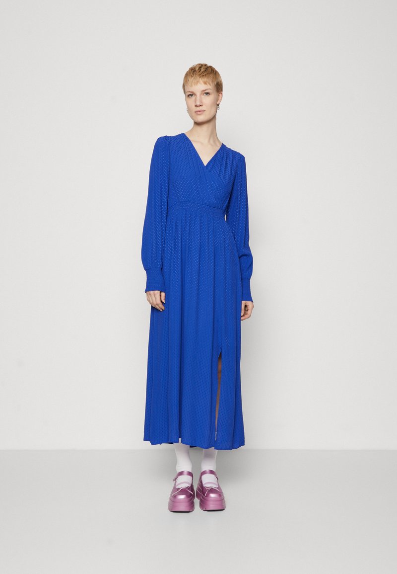 Abito blu a maniche lunghe con design avvolgente, tessuto testurizzato e uno spacco fino alla coscia. Indossato con calzini bianchi e scarpe con piattaforma rosa.