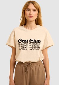 T-shirt en coton beige à manches courtes, présentant un texte graphique noir "Cool Club" dans une police stylisée. Associé à un pantalon à cordon de serrage marron.