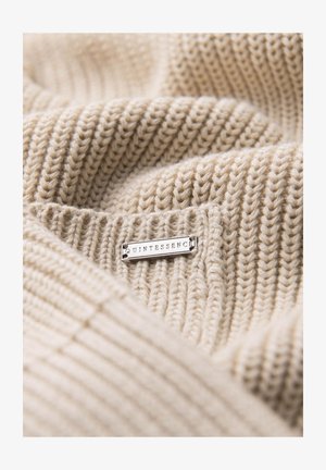 Beżowa dzianina z wyraźnym wzorem prążkowym, z dołączoną srebrną metką z napisem "QUINTESSENCE". Materiał wydaje się miękki i przytulny.