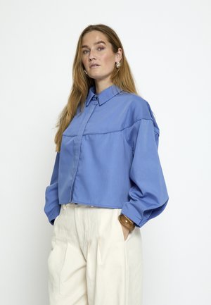 Femme aux cheveux longs portant une chemise bleue ample à boutons et un pantalon plissé crème, debout les mains dans les poches devant un fond uni.