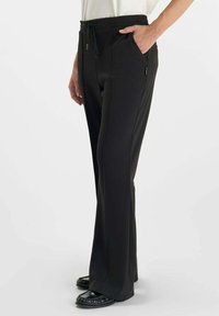 Vrouw in zwarte recht-door-de-zaak broek met zijzakken en zwarte loafers, staand tegen een effen witte achtergrond.