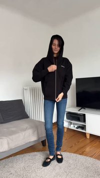 Svart hettejakke med glidelås, stor hette, frontlommer og logo. Kombinert med blå tettsittende jeans og svarte flate sko. Rommiljø.