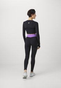 Top preto de mangas longas com design cropped, combinado com leggings pretas com uma faixa roxa na cintura. Modelo está de costas, usando tênis brancos.