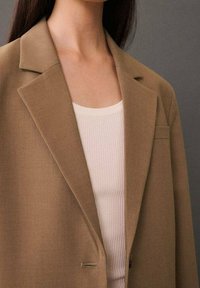 Femme portant un blazer beige sur un débardeur blanc côtelé, sur un fond gris uni.