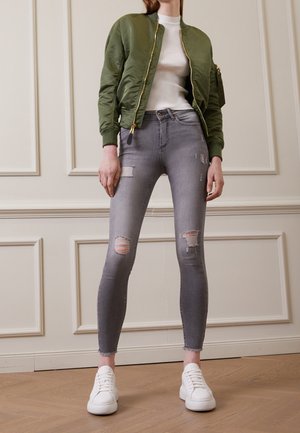 Person trägt grüne Bomberjacke, weißes eng anliegendes Top, zerrissene graue Skinny-Jeans und weiße Turnschuhe, steht drinnen vor einer getäfelten Wand.