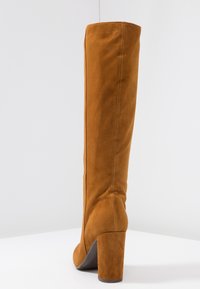 Bota alta de ante para mujer en color marrón con un tacón alto y grueso, mostrada desde atrás sobre una superficie blanca y fondo claro.