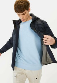 Navy jacket with quilted texture, zippered front, adjustable hood, and gray lining, worn over a light blue sweatshirt and cream cargo pants.