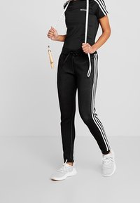 Top athlético negro con rayas blancas y logo, combinado con pantalones deportivos texturizados negros con rayas blancas en los costados y cintura atada, rematado con zapatillas grises.