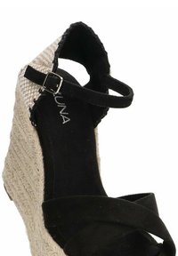 Sandalia de cuña de ante negro con una base de esparto, presenta un diseño trenzado en la parte superior y una correa ajustable en el tobillo.