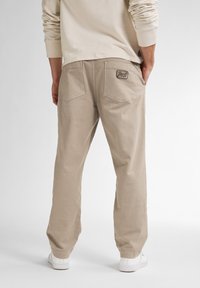 Person som bär beige avslappnade byxor med bakfickor, där en har en "Petrol"-brandlapp, i kombination med en ljusbeige långärmad skjorta och vita sneakers.