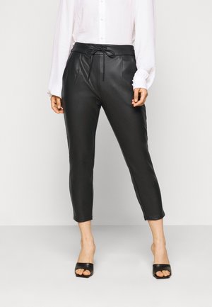 Sorte, kunstlæderbukser med indsnævret pasform, snøre i taljen og forplisseringer foran. Kombineret med en hvid skjorte med knapper og sorte slides.