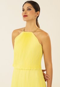 Gelbes, plissiertes Kleid mit Halterneck, verziert mit goldfarbenen Kettenakzenten. Der Stoff hat eine glatte Textur und eine fließende Silhouette.
