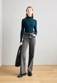 Teal tettsik med høy hals og striper, kombinert med høytlivede grå brede bukser. Modellen bærer en svart plissépåse og har på seg svarte sko.