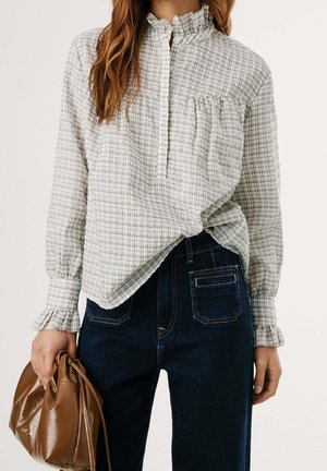 Femme portant un chemisier blanc à carreaux avec un col et des poignets à volants, un jean bleu foncé, tenant un sac à main en cuir marron.